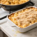 Baked macaroni and cheese in a white ceramic dish with a cheesy skillet in the background