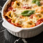 Creamy tomato basil tortellini bake in a baking dish on a rustic wooden table, surrounded by plates, red wine, garlic bread, and a salad.