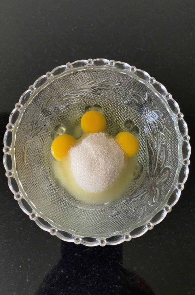 Close-up of eggs, sugar, salt, and vanilla extract being added to a mixing bowl for an Almond Caramel Layer Cake.