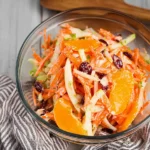 Bowl of vibrant carrot apple salad with mandarin slices, cranberries, and creamy dressing, served chilled on a dark ceramic plate.