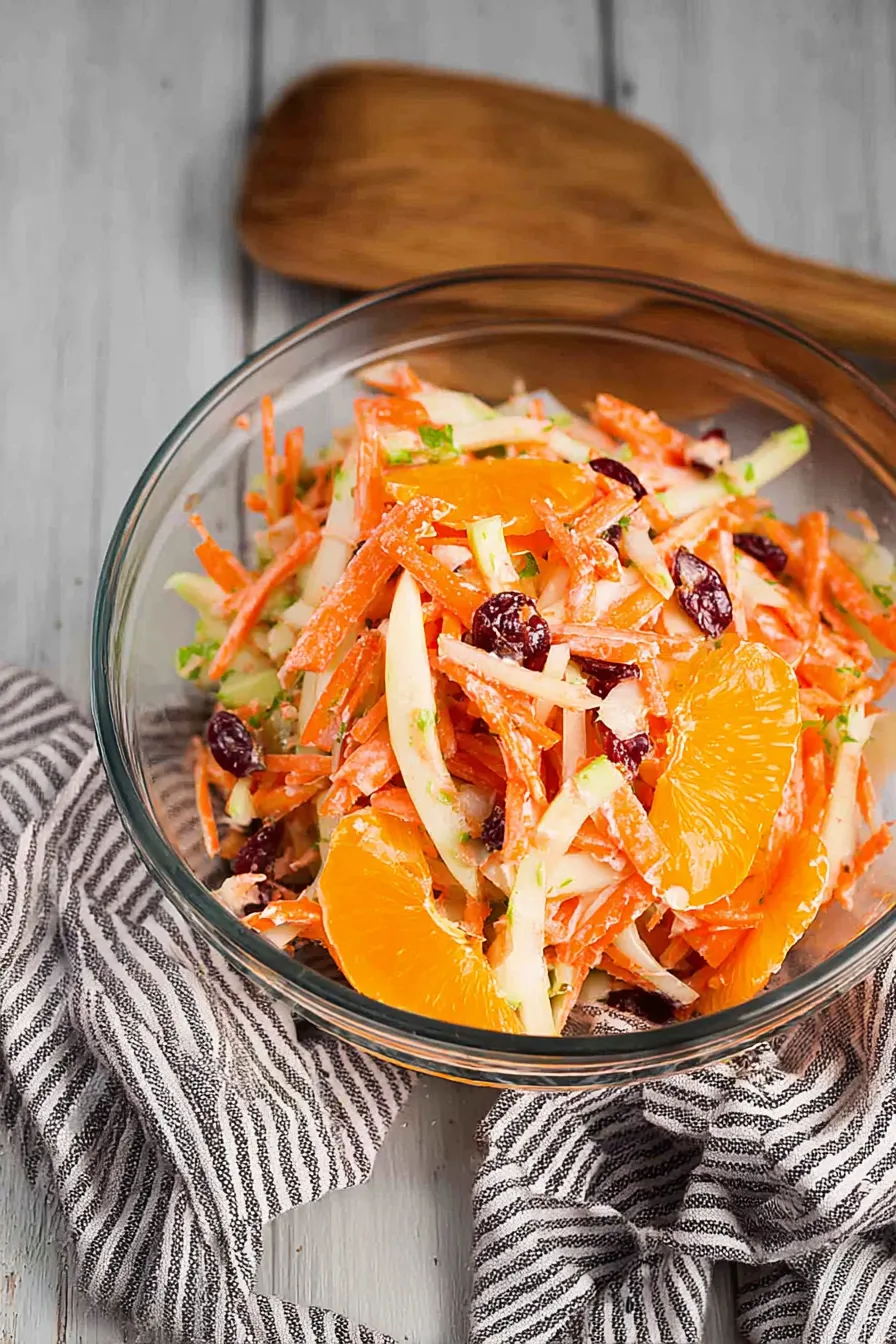 Bowl of vibrant carrot apple salad with mandarin slices, cranberries, and creamy dressing, served chilled on a dark ceramic plate.