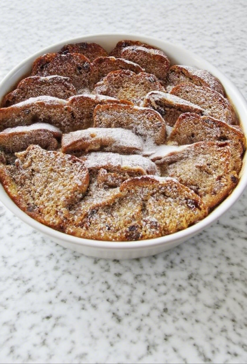 Hot cross bun pudding fresh from the oven, dusted with icing sugar on a white kitchen counter.