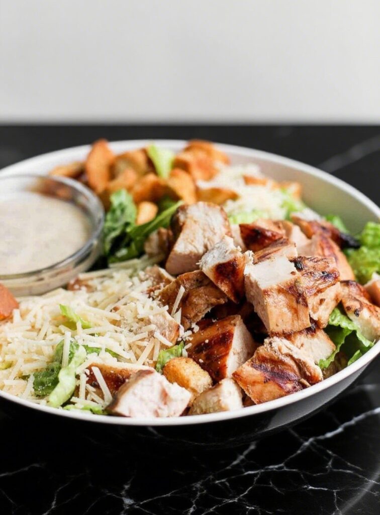 Sliced grilled chicken breasts served on a white plate with Caesar Salad, croutons, parmesan cheese, and Caesar dressing.