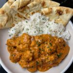 Delicious Butter Chicken with tender chicken chunks in a rich orange sauce, garnished with parsley, served with naan and rice.
