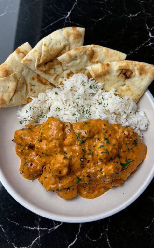 Delicious Butter Chicken with tender chicken chunks in a rich orange sauce, garnished with parsley, served with naan and rice.