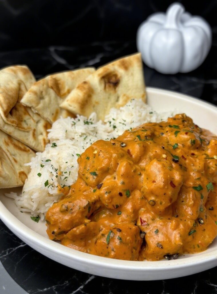 Butter chicken with creamy sauce, served with naan bread and rice, garnished with parsley, showcasing vibrant orange and white textures.