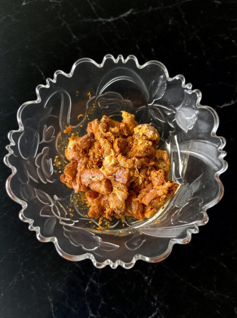 Close-up of bite-sized boneless chicken pieces marinated in a vibrant orange-red mixture with visible spice flecks.